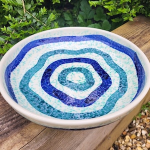 May include: A white ceramic bowl with a blue and green concentric circle design. The bowl has a speckled glaze and a slightly wavy rim.