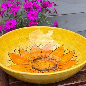 May include: A large, round, yellow ceramic bowl featuring a hand-painted sunflower design in orange and black. The bowl is placed on a wooden surface, with vibrant pink flowers in the background. This decorative bowl is perfect for serving or display.