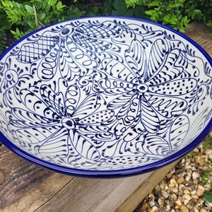 May include: Oval ceramic serving bowl with a white base and intricate blue floral patterns. The rim and outer edge are a solid blue. The bowl is placed on a wooden surface, with green foliage in the background.