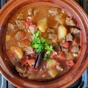 May include: A close-up of a terracotta pot filled with a stew of beef, potatoes, tomatoes, and herbs. The stew is simmering in a rich, brown sauce and garnished with fresh parsley.