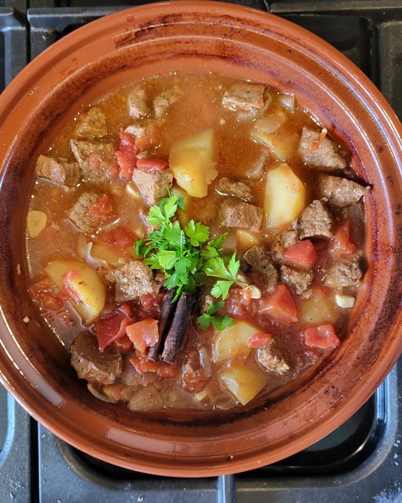 May include: A close-up of a terracotta pot filled with a stew of beef, potatoes, tomatoes, and herbs. The stew is simmering in a rich, brown sauce and garnished with fresh parsley.