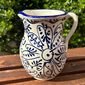 May include: A white ceramic jug with a blue floral pattern. The jug features a curved handle and a spout with a blue rim. The floral design is hand-painted in blue and covers the body of the jug.