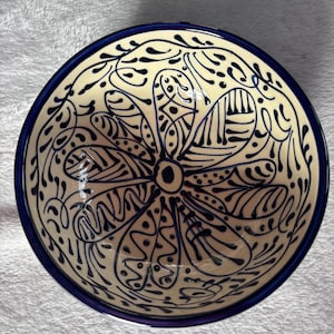 May include: A cream-coloured ceramic bowl with a dark blue rim. The bowl has a detailed floral pattern in dark blue, featuring a central flower design and intricate leaf and dot motifs. The bowl is likely used for serving food.