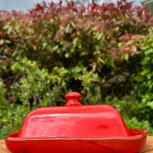 May include: A red ceramic butter dish with a lid. The dish is sitting on a wooden surface.
