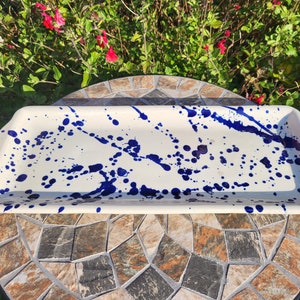 May include: A rectangular ceramic serving tray with a smooth off-white base featuring a vibrant cobalt blue paint splatter pattern across the surface. The tray has slightly raised edges and integrated handles on both ends for easy carrying.