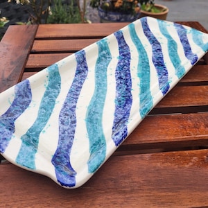 May include: A white ceramic serving tray with blue and turquoise wavy stripes. The tray is rectangular and has a slightly curved edge.