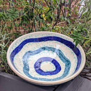 May include: A ceramic bowl with a white interior and exterior. The bowl features concentric, wavy bands of blue and teal, creating a spiral design. The exterior has a speckled pattern. The bowl is round and appears to be handmade.