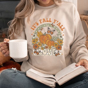 Pode incluir: Uma mulher vestindo uma camiseta bege com o texto "It's Fall Y'all Love" em uma fonte de estilo vintage. A camiseta apresenta um gr&aacute;fico de ab&oacute;boras, borboletas e flores. Ela est&aacute; segurando uma caneca branca e um livro.