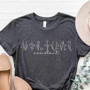 May include: A dark gray t-shirt with the text "VIRTUAL assistant" in white lettering with floral accents. The shirt is laid flat on a white surface with a pair of beige flats and a straw hat.