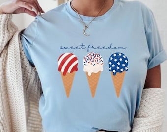May include: A light blue t-shirt with a graphic of three ice cream cones decorated with red, white, and blue. The text "sweet freedom" is printed on the shirt.