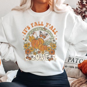 Pode incluir: Camisola branca com um design gr&aacute;fico que apresenta ab&oacute;boras, borboletas e o texto "It's Fall Y'all Love".