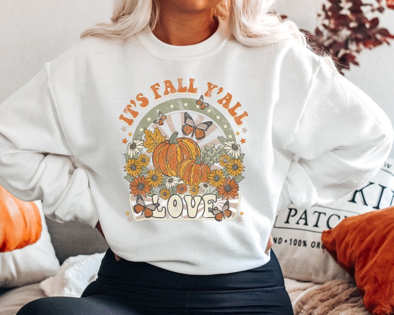 Pode incluir: Camisola branca com um design gr&aacute;fico que apresenta ab&oacute;boras, borboletas e o texto "It's Fall Y'all Love".