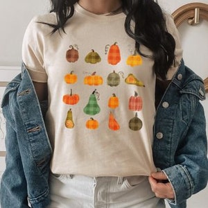 May include: A beige t-shirt featuring a graphic print of assorted pumpkins in shades of orange, green, and brown. The pumpkins are arranged in a grid pattern and come in various shapes and sizes.