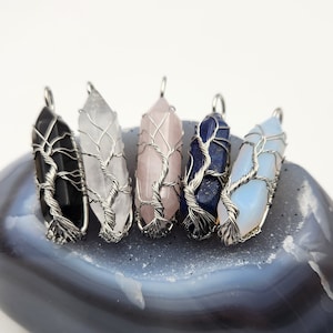 May include: Five wire-wrapped crystal pendants in various colours, including black, white, pink, blue, and light blue, are displayed on a grey stone surface.
