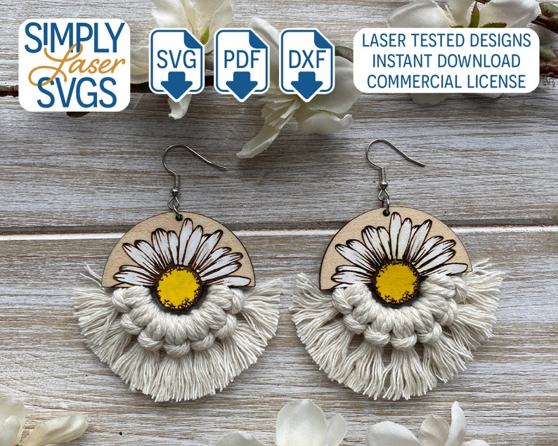 May include: A pair of wooden earrings with a white daisy design and white macrame fringe. The earrings are on a wooden surface.