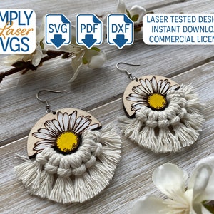 May include: A pair of wooden earrings with a white daisy design and white macrame fringe. The earrings are on a wooden surface.