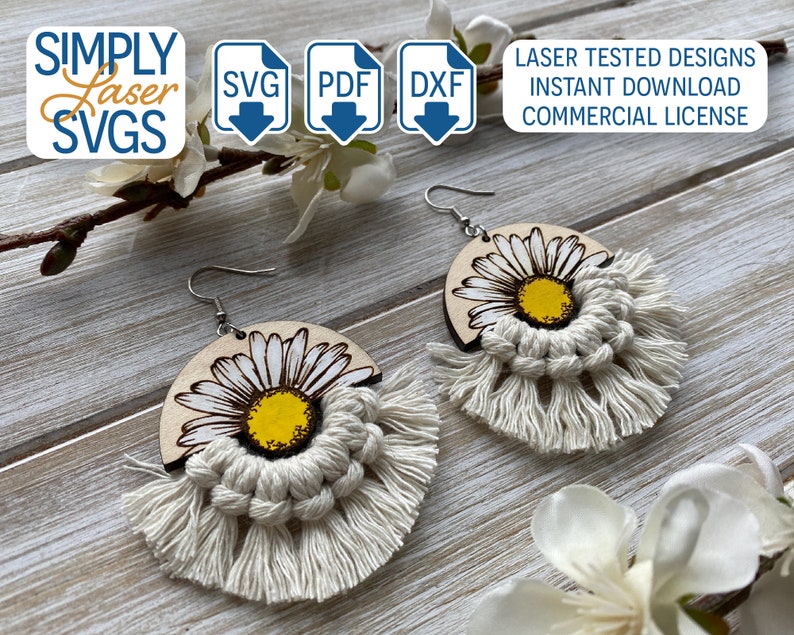 May include: A pair of wooden earrings with a white daisy design and white macrame fringe. The earrings are on a wooden surface.