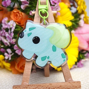 Op de afbeelding: Een lichtblauwe en groene cartoonfiguur sleutelhanger met glitter. De sleutelhanger is bevestigd aan een groene metalen sluiting en wordt getoond op een houten ezel.