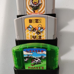 May include: Three Nintendo 64 game cartridges in a black plastic holder. The top cartridge is gray and has a yellow daisy with a smiley face on it. The text on the cartridge reads 'Daisy Distortion'. The middle cartridge is gray and has a yellow honeycomb pattern with three bees on it. The text on the cartridge reads 'Buzz Fuzz'. The bottom cartridge is green and has a blue sky with white clouds and a cow in a field. The text on the cartridge reads 'Grazer Phaser'. 