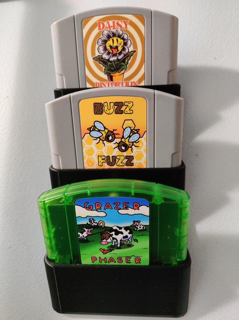 May include: Three Nintendo 64 game cartridges in a black plastic holder. The top cartridge is gray and has a yellow daisy with a smiley face on it. The text on the cartridge reads 'Daisy Distortion'. The middle cartridge is gray and has a yellow honeycomb pattern with three bees on it. The text on the cartridge reads 'Buzz Fuzz'. The bottom cartridge is green and has a blue sky with white clouds and a cow in a field. The text on the cartridge reads 'Grazer Phaser'. 