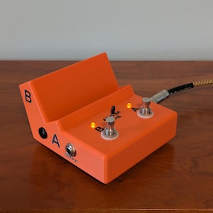 May include: An orange guitar effects pedal featuring two metal footswitches labelled A and B, two illuminated amber indicator lights, and a central toggle switch. The unit includes an output jack, power input, and side-mounted labels A and B.
