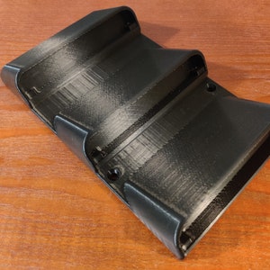 May include: A black plastic 3D printed wall mount with three slots for holding items. The slots are angled slightly upward and have a smooth, rounded edge.