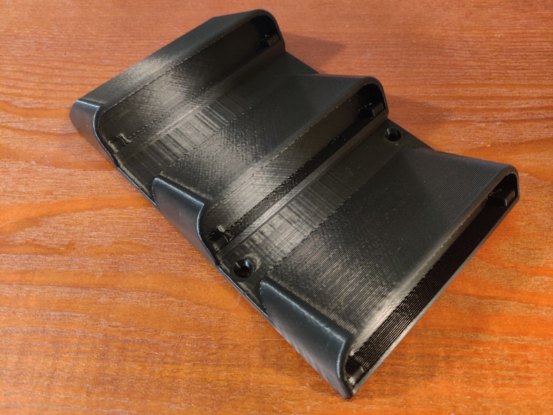 May include: A black plastic 3D printed wall mount with three slots for holding items. The slots are angled slightly upward and have a smooth, rounded edge.