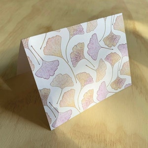 May include: A folded greeting card with a white background and a pattern of stylized ginkgo leaves in shades of purple, peach, and gold. The card is propped up on a light-colored wooden surface.