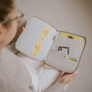 Könnte beinhalten: Hellgraue, mit Reißverschluss versehene Organizer-Geldbörse mit mehreren Fächern. Die Geldbörse ist geöffnet und zeigt gelbe Karten und andere Dokumente. Der Innenraum umfasst Kartenfächer und eine Reißverschlusstasche. Der Reißverschluss und die Beschläge sind goldfarben.