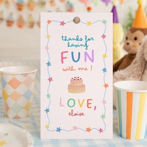May include: A rectangular white paper gift tag with a hole at the top, featuring a light blue border with colourful stars. The text reads: thanks for having FUN with me! LOVE, eloise. A small illustration of a two-tier pink birthday cake is centred.