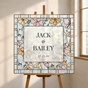 Könnte beinhalten: Ein Hochzeitszeichen aus Glasmalerei auf einer Staffelei. Das Schild zeigt die Namen "Jack & Bailey" und das Datum "07.25.26". Es ist mit rosa, gelben und orangen Blumen und grünen Ranken verziert. Der Hintergrund ist hellblau.