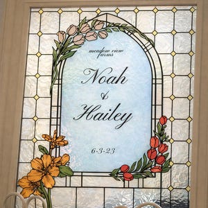 Può includere: Un pannello decorativo in vetro colorato con i nomi "Noah & Hailey" e la data "6.3.23". Il design include elementi floreali arancioni, rosa e rossi, su sfondo azzurro chiaro, incorniciato in una cornice color crema.