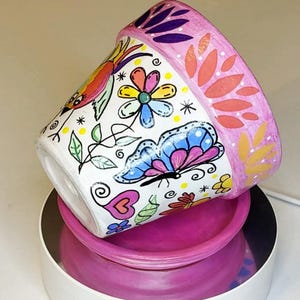 Könnte beinhalten: Ein weißer Keramik-Pflanztopf mit handgemaltem Design, das einen bunten Schmetterling, eine mehrfarbige Blume, ein Herz und einen Vogel zeigt. Der Rand ist rosa bemalt mit orangefarbenen und gelben Blattmustern und steht auf einem rosa Untersetzer.