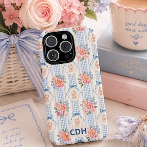May include: A phone case featuring a pattern of blue and white gingham stripes, pink floral bouquets, and small teddy bears. The case includes the letters CDH printed in dark blue on the lower portion of the back surface.