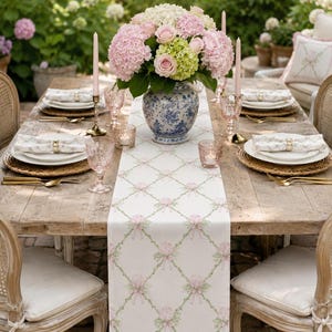 May include: A white table runner featuring a green lattice pattern with pink floral accents and bows, centered on a wooden dining table set with white plates, gold cutlery, pink glassware, tall taper candles, and a blue and white floral vase.