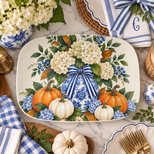 Puede incluir: Una bandeja de servicio ovalada de cerámica con un jarrón floral azul y blanco en el centro, lleno de flores de hortensia color crema y bayas azules, acentuado por un lazo a rayas azul y blanco, flanqueado por calabazas naranjas y blancas.