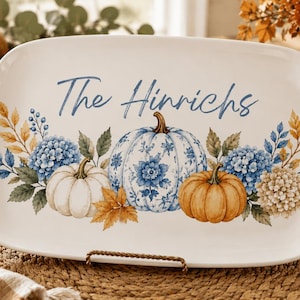 Puede incluir: Una bandeja de servicio rectangular de cerámica blanca con el texto en azul The Hinrichs en el centro, rodeado por una ilustración de tres calabazas con patrones florales en naranja, blanco y azul, decorada con hortensias azules y beige.