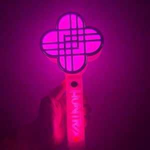 Puede incluir: Un palo de luz rosa brillante con una parte superior en forma de trébol de cuatro hojas y la palabra "huntra" impresa en el mango. El palo de luz emite un brillo rosa brillante, sobre un fondo oscuro.