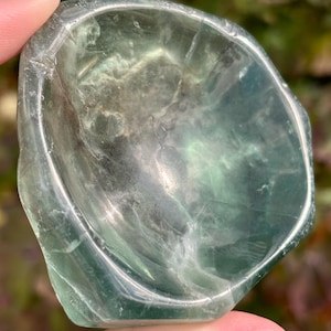 May include: A polished, bowl-shaped object carved from translucent green fluorite mineral. The stone features natural internal veining, cloudy white inclusions, and varying shades of light to dark green throughout its smooth, concave surface.