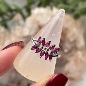 May include: A silver ring with a cluster of marquise-cut, deep pink gemstones. The ring's design resembles a floral arrangement, with small clear stones accenting the main cluster. The ring is displayed on a light pink, cone-shaped object.