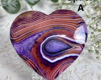 Peut inclure: Une pierre d'agate polie en forme de cœur présentant des bandes concentriques violettes, orange brûlé et blanches, avec une cavité géode cristalline centrale. La pierre est posée sur un support en plastique transparent, marquée Option A.