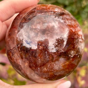 May include: A polished spherical crystal featuring deep red and burnt orange inclusions within a translucent, clear quartz base. The surface is smooth and reflective, showcasing internal fractures and natural mineral patterns throughout the orb.