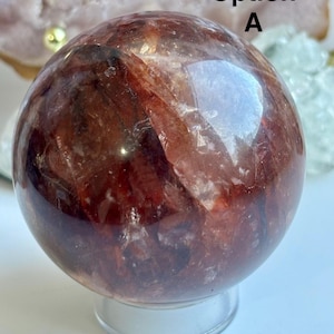 Peut inclure: Une grande sphère de cristal polie, de couleur brun-rouge, reposant sur un support clair et circulaire. La surface de la sphère reflète la lumière, révélant des motifs internes. Le texte "Option A" est visible. Il s'agit d'une boule de cristal décorative.