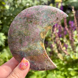 May include: A crescent moon-shaped stone carving featuring a mottled surface with a blend of deep pink, forest green, and earthy brown mineral inclusions. The polished stone displays natural variations in colour and texture across its curved form.
