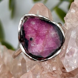 Op de afbeelding: Een zilveren ring met een grote, onregelmatige, ruwe plak roze toermalijn. De steen heeft een levendig magenta centrum met donkere, bijna zwarte randen, gezet in een gladde, gepolijste zilveren metalen band die de organische vorm volgt.
