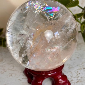 May include: A clear, spherical quartz crystal ball featuring internal fractures, natural inclusions, and a vibrant rainbow iridescence near the top. The sphere rests securely on a dark red, carved wooden stand with a circular base and curved legs.