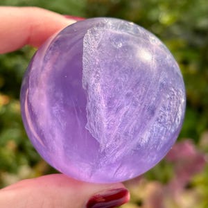 May include: A polished, spherical amethyst crystal featuring a translucent light purple hue. The stone displays natural internal inclusions and veining patterns that create a textured, cloud-like appearance within the smooth, rounded surface.