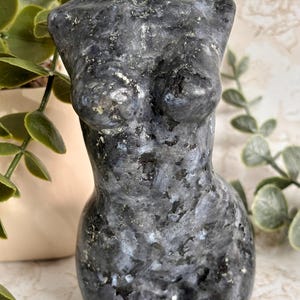 Pode incluir: Uma escultura de pedra labradorita polida esculpida na forma de um torso humano. A pedra apresenta uma cor base cinza-escura com manchas iridescentes azuis e prateadas por toda a superfície, exibindo a textura mineral natural da peça.