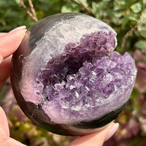 May include: A spherical amethyst geode featuring a polished dark exterior and a hollow centre filled with deep purple, jagged crystalline structures. The natural stone formation displays a contrast between the smooth outer surface and the rough, sparkling interior.
