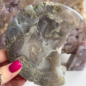 May include: A polished, crescent-shaped stone with a marbled pattern in shades of grey, white, and tan. The stone is held in a hand with a bright pink fingernail. Other stones in purple and brown are in the background.